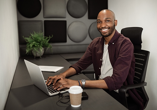 man sitting at his desk with an open laptop and a takeaway cup of coffee
