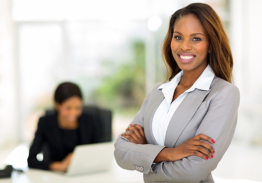 woman standing with her arms cross with another woman in the background sitting at her desk working on her laptop